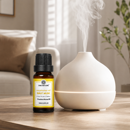 A white diffuser and a small amber bottle of essential oil sit on a wooden table in a softly lit living room. The diffuser is emitting a visible plume of steam, and the bottle label reads One Nature Brut Musk.