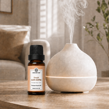 A small amber bottle of One Nature Fresh Cape Coffee aroma oil stands beside a matte cream-colored diffuser on a wooden table, with steam rising from the diffuser in a softly lit living room setting.