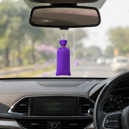 A purple car air freshener hanging ornament is suspended from the rearview mirror inside a car, with blurred trees and other vehicles visible through the windshield.