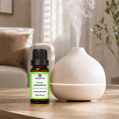 A small amber bottle of One Nature Lively Lemongrass essential oil stands beside a white ceramic diffuser on a wooden table in a softly lit living room. Steam rises from the diffuser, and the product label is clearly visible.