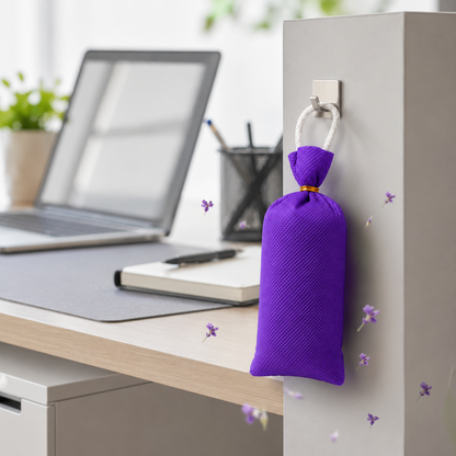 A purple textured fabric hanging bag with a white loop hangs from a wall hook on a light gray office cubicle wall, with a blurred desk scene including a laptop and notebook in the background.