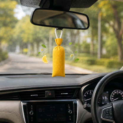 A yellow fabric car air freshener bag hanging from the rearview mirror inside a car, with a blurred tree-lined road visible through the windshield.